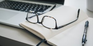 Computer, glasses, notebook, and pen at a work station.